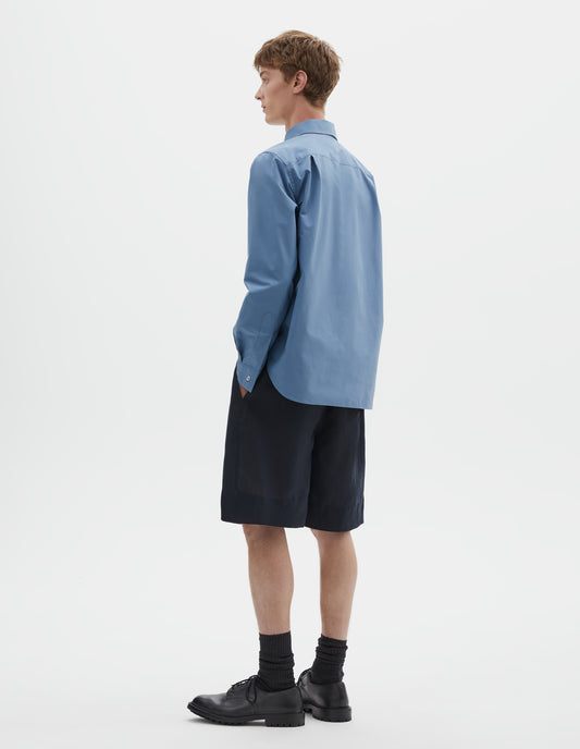 Full body image, back view of model wearing blue cotton long sleeve shirt, with mother of pearl button cuffs and pleats on back yoke. Styled with dark navy blue shorts, black socks and black leather brogues.