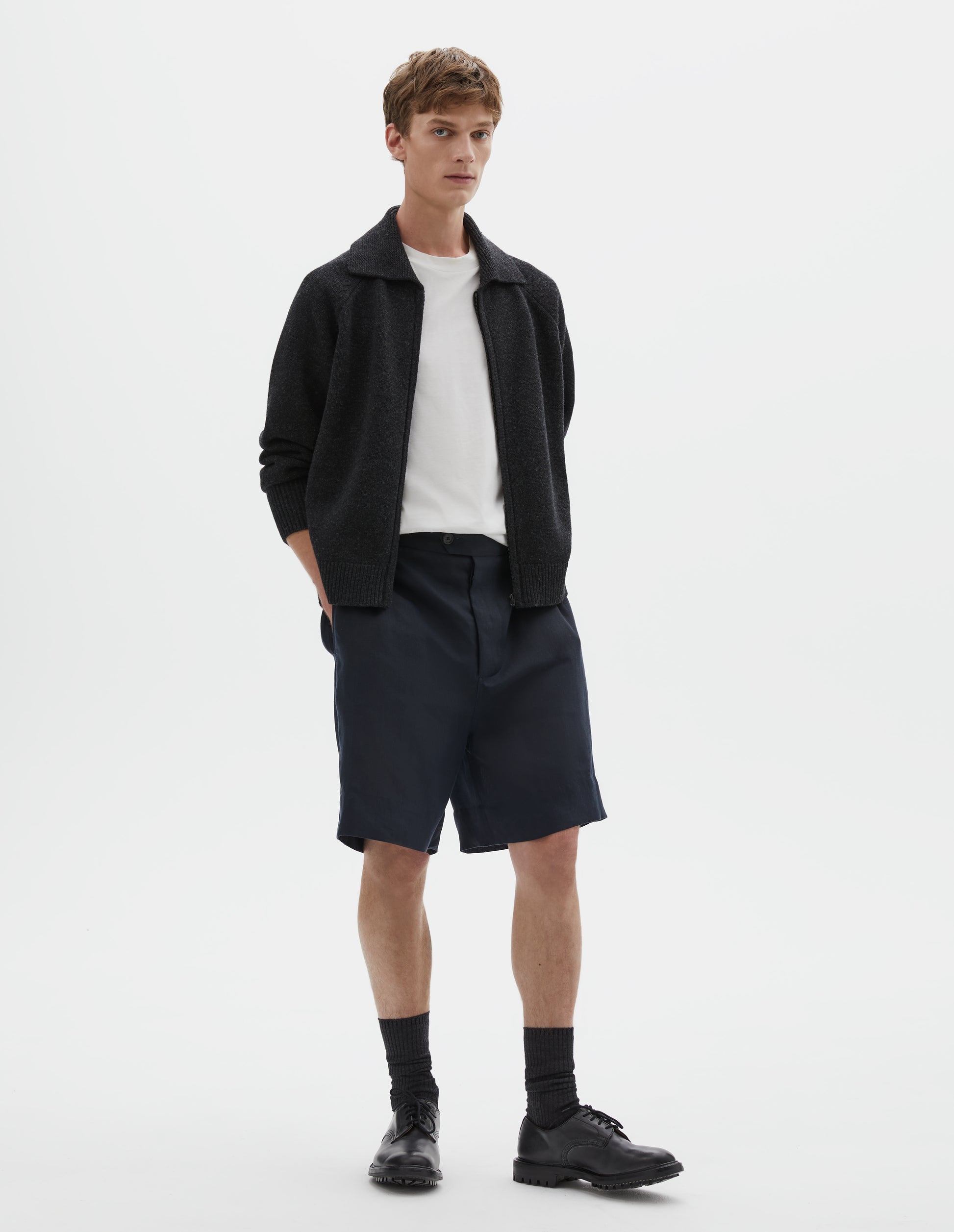 Full body image, front view of model wearing relaxed, knee length dark blue shorts with half-elasticated waistband with belt loops, horn button fly with button tab fastening and slant side pockets. Styled with dark grey cardigan and white t-shirt, black socks and black leather shoes.