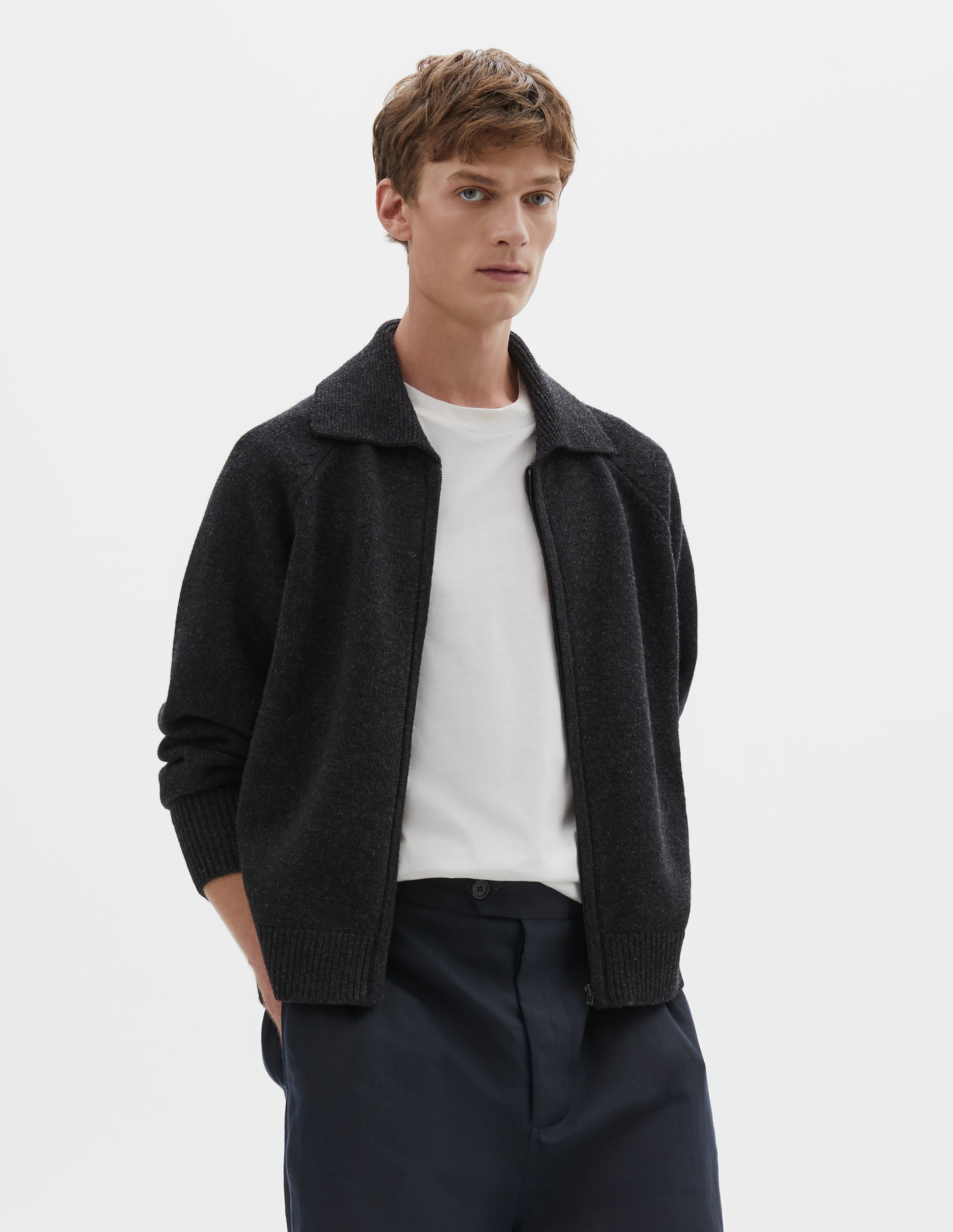 Cropped body image, front view of model wearing dark grey wool cardigan, with raglan sleeve, zip fastening and contrast rib on collar, cuffs and hem. Styled over a white t-shirt, with dark navy blue shorts.  