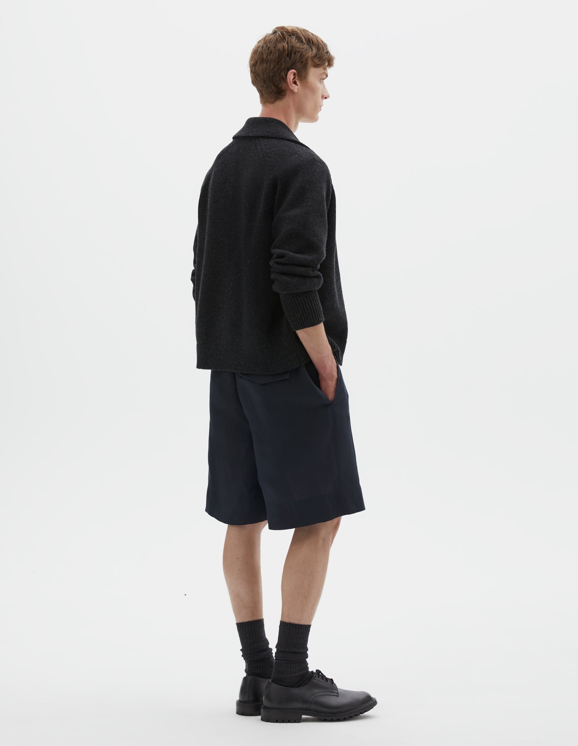 Full body image, back view of model wearing relaxed, knee length dark blue shorts with half-elasticated waistband with belt loops, slant side pockets, and welt and concealed horn button-fastening flap back pockets. Styled with dark grey cardigan, black socks and black leather shoes.