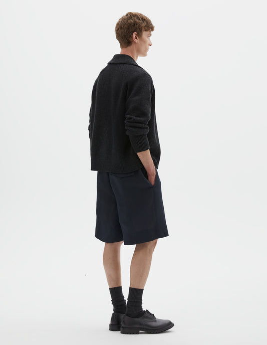 Full body image, back view of model wearing relaxed, knee length dark blue shorts with half-elasticated waistband with belt loops, slant side pockets, and welt and concealed horn button-fastening flap back pockets. Styled with dark grey cardigan, black socks and black leather shoes.