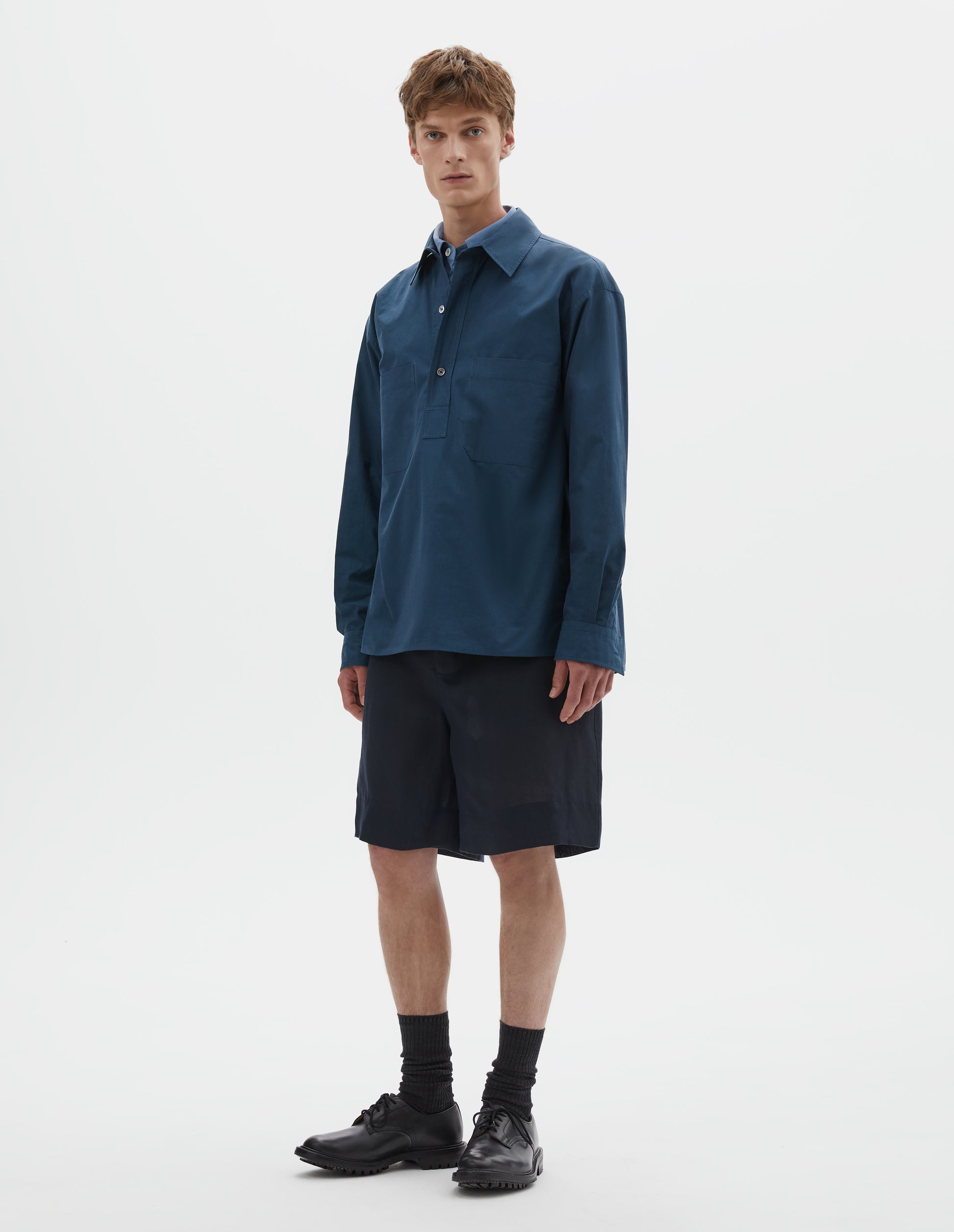 Full body image, front view of model wearing relaxed blue shirt with mother of pearl buttons on stitch through raised placket, front patch pockets and deep side split vents. Styled over light blue shirt, with dark blue shorts and black socks with black leather shoes.