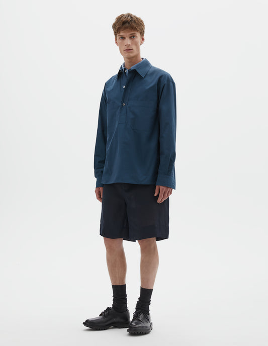 Full body image, front view of model wearing relaxed blue shirt with mother of pearl buttons on stitch through raised placket, front patch pockets and deep side split vents. Styled over light blue shirt, with dark blue shorts and black socks with black leather shoes.