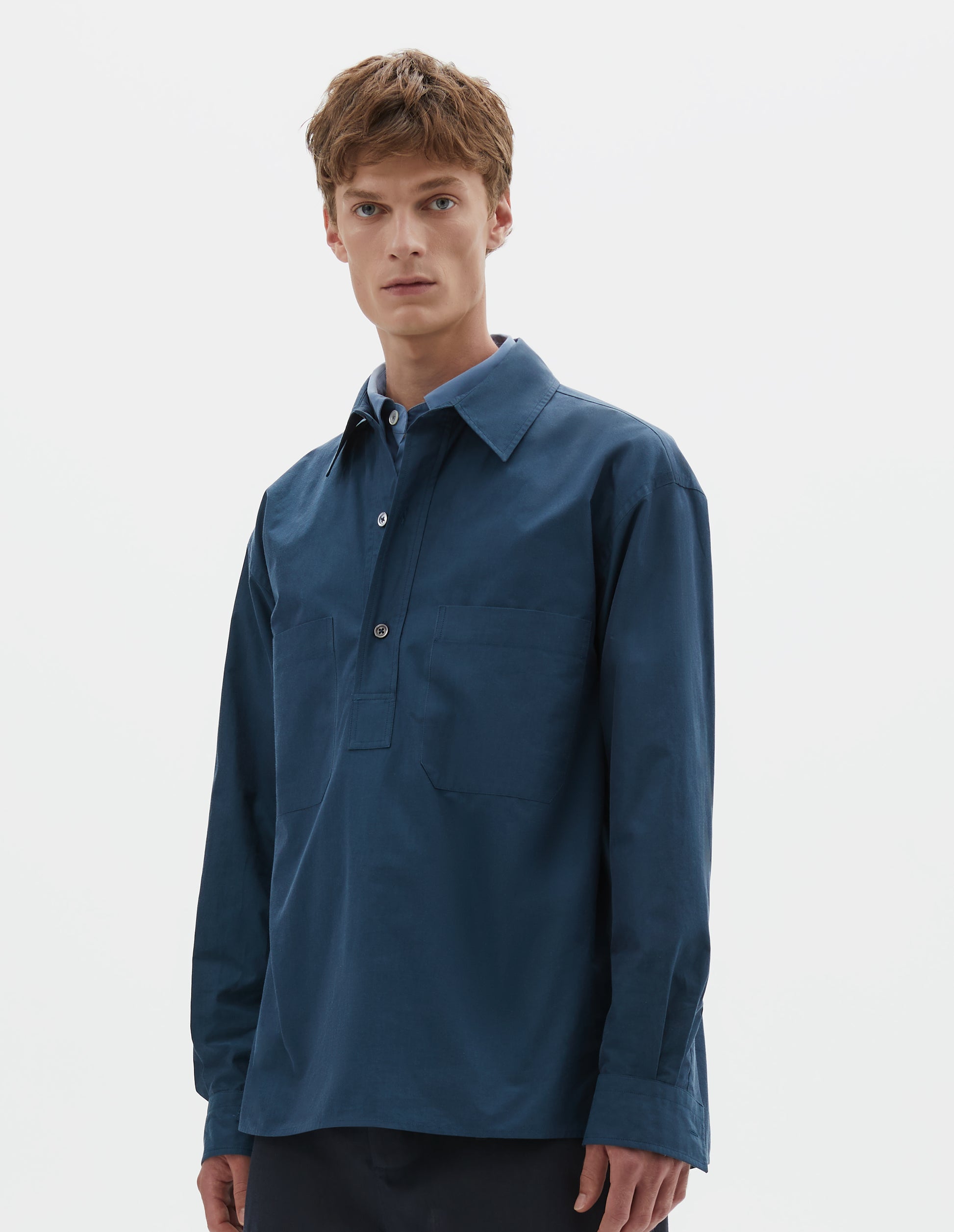 Cropped body image, front view of model wearing relaxed blue shirt with  mother of pearl buttons on stitch through raised placket, front patch pockets and deep side split vents. Styled over light blue shirt and with dark blue shorts.