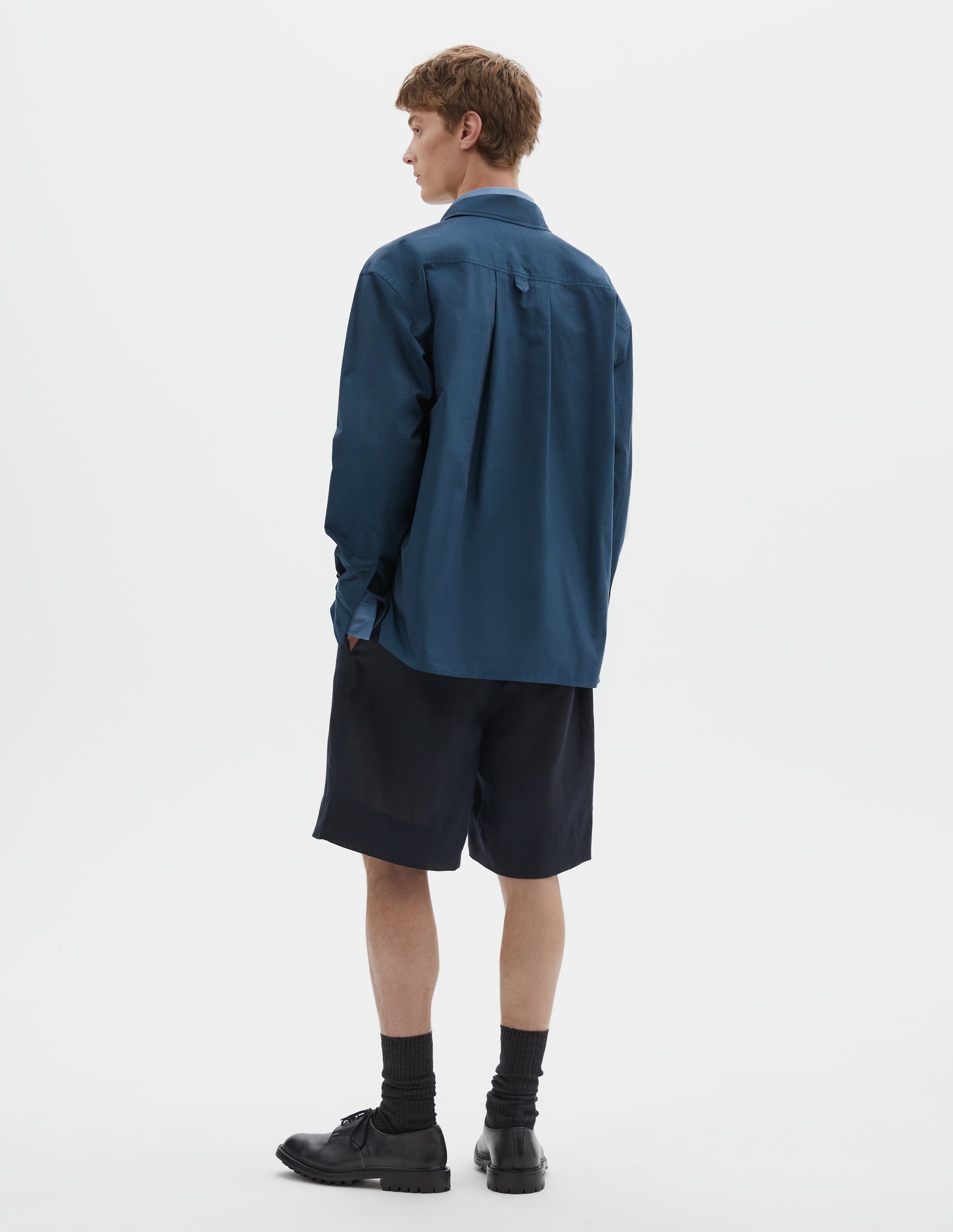 Full body image, back view of model wearing relaxed blue shirt with deep side split vents, elongated back, box pleat and hanger loop detail on back yoke. Styled over light blue shirt, dark blue shorts and black socks with black leather shoes.