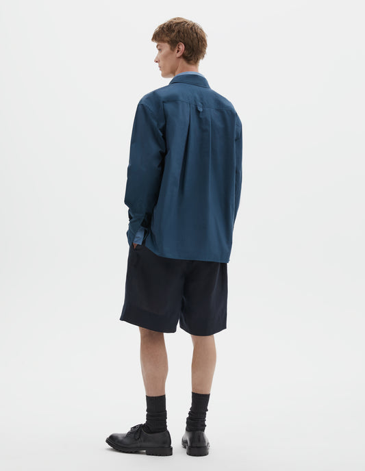Full body image, back view of model wearing relaxed blue shirt with deep side split vents, elongated back, box pleat and hanger loop detail on back yoke. Styled over light blue shirt, dark blue shorts and black socks with black leather shoes.
