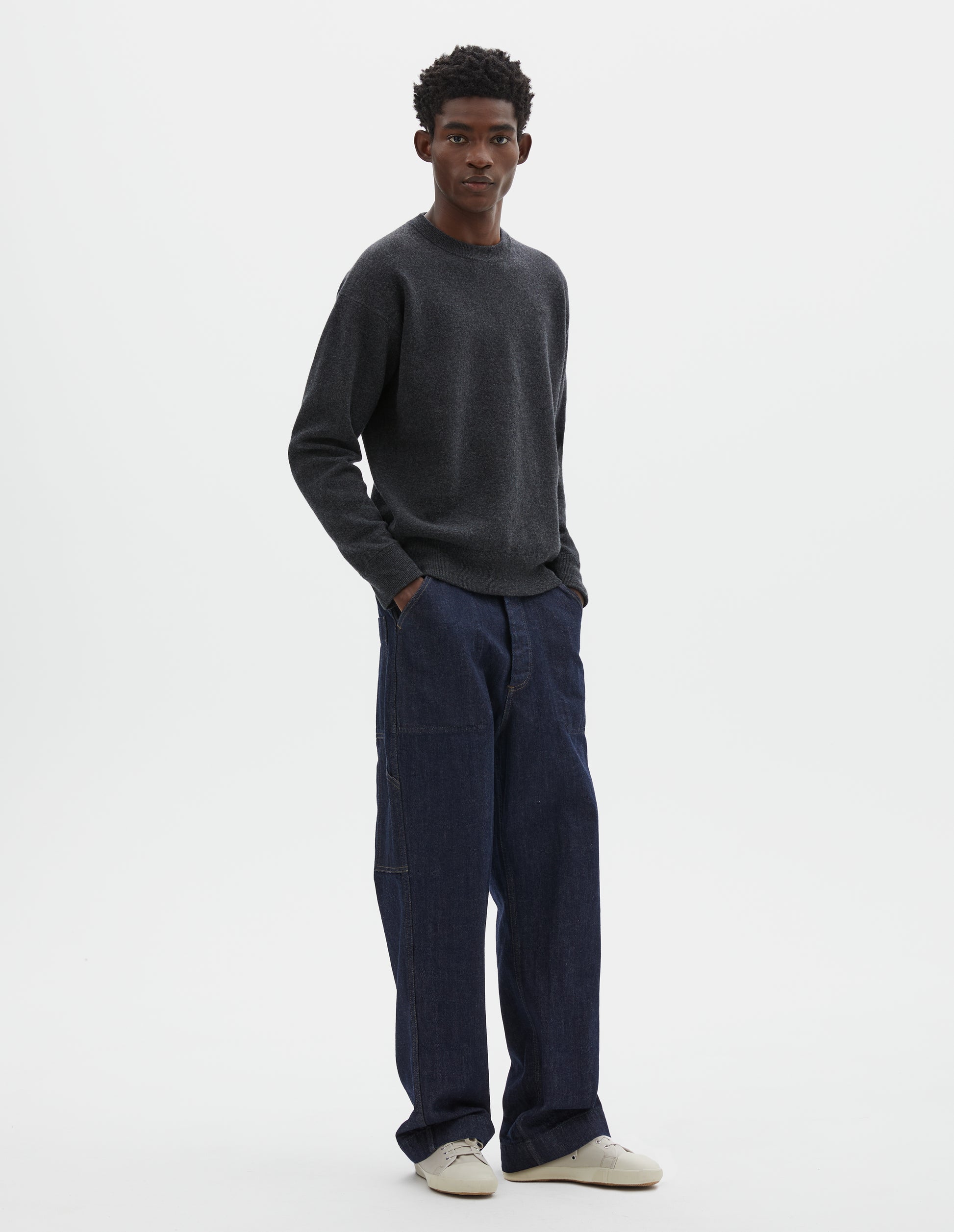Full body image, front view of model wearing relaxed  dark grey knitted sweatshirt with rib collar, cuffs and hem. Styled with dark blue jeans and beige trainers.