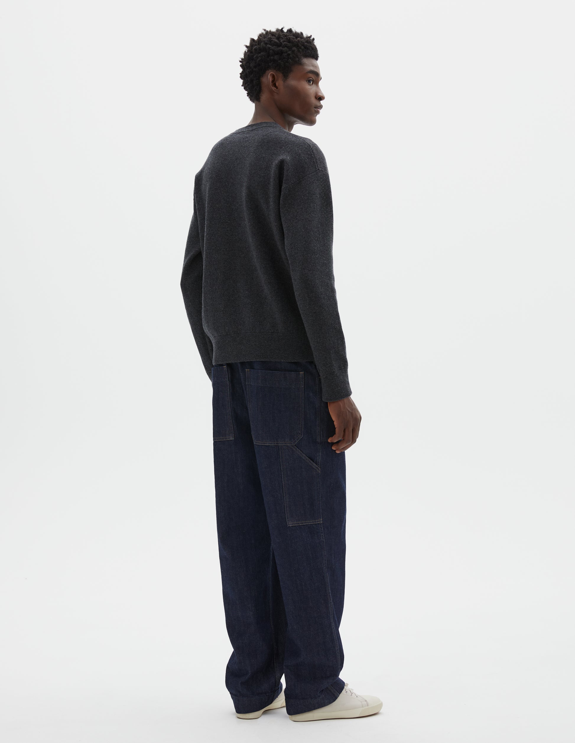 Full body image, back view of model wearing relaxed  dark grey knitted sweatshirt with rib collar, cuffs and hem. Styled with dark blue jeans and beige trainers.