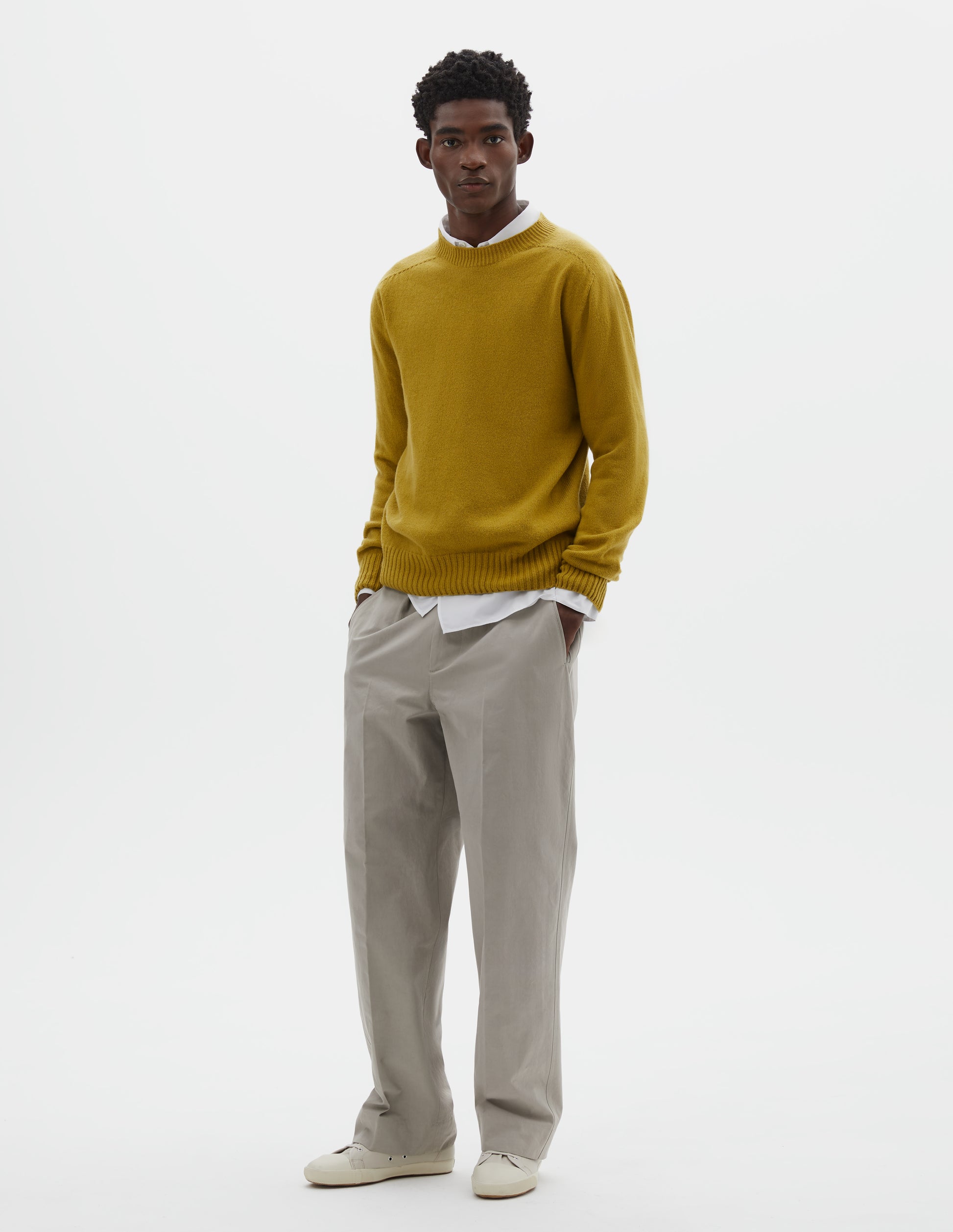 Full body image, front view of model wearing yellow crew neck jumper with seamless finish and contrast ribs on neck, cuffs and hem. Styled over white cotton shirt, with grey trousers and off white canvas trainers.