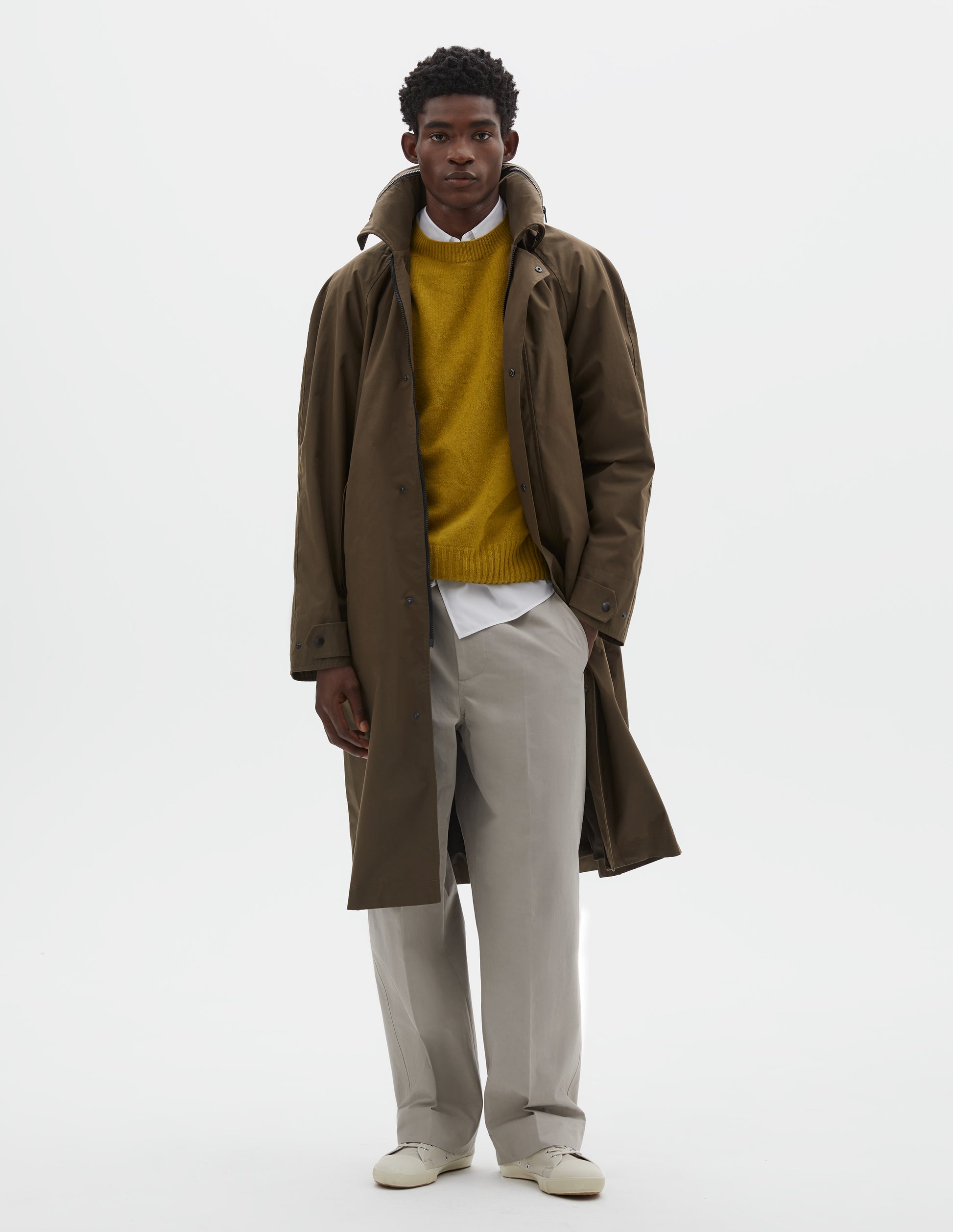 Full body image, front view of model wearing greenish brown ventile cotton poplin longline mac, with slant pockets, vislon zip and adjustable popper cuffs. Styled over white shirt, yellow jumper, beige trousers and off white canvas trainers.