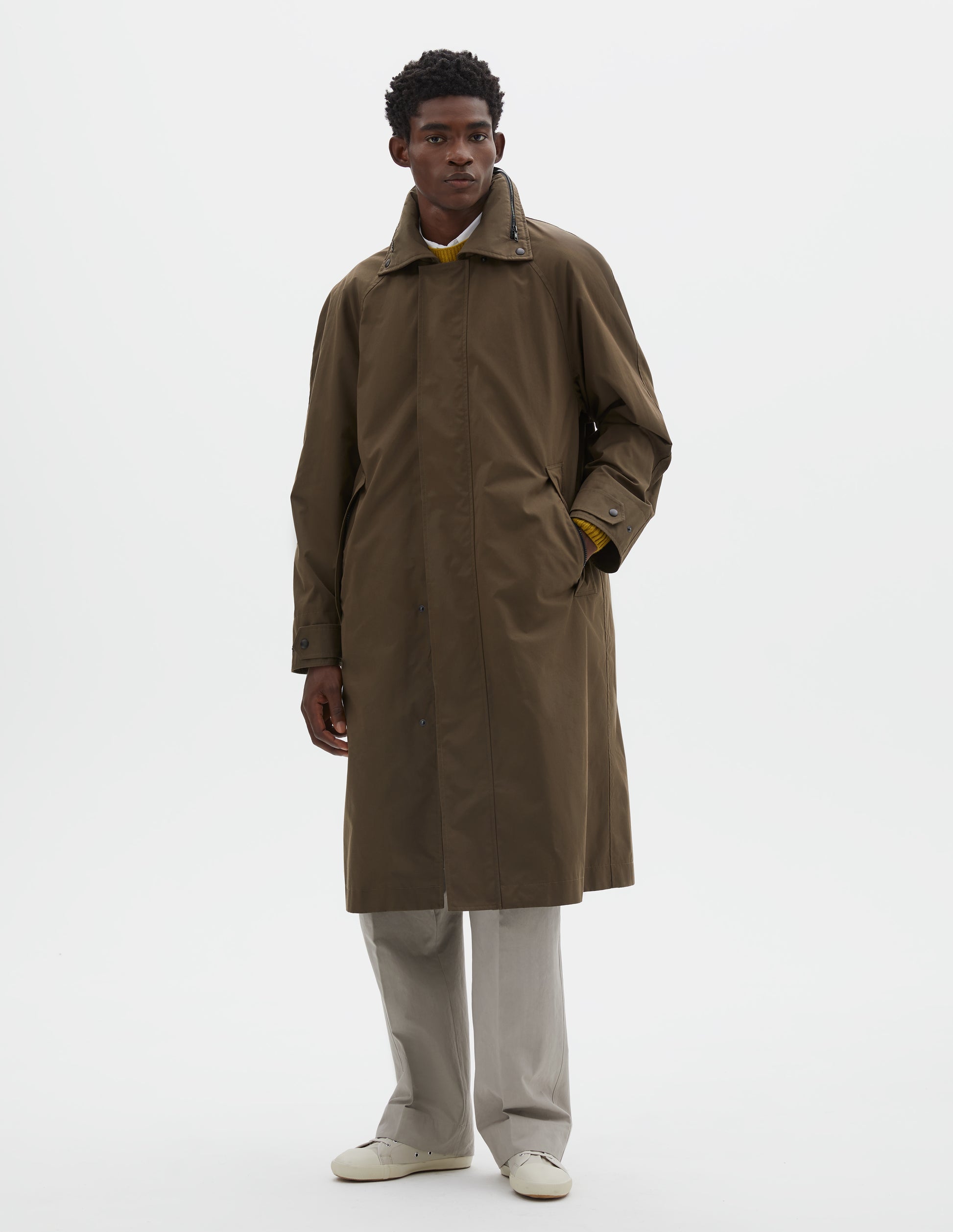Full body image, front view of model wearing greenish brown ventile cotton poplin longline mac, with slant pockets, adjustable popper cuffs and packable hood. Styled over white shirt, yellow jumper, beige trousers and off white canvas trainers.