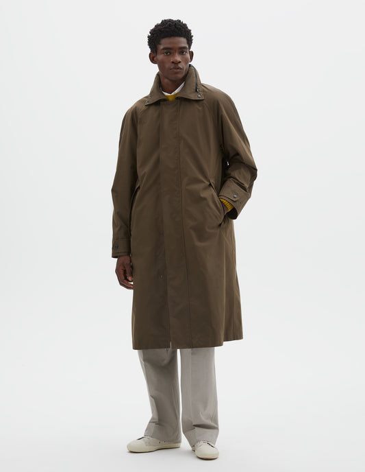 Full body image, front view of model wearing greenish brown ventile cotton poplin longline mac, with slant pockets, adjustable popper cuffs and packable hood. Styled over white shirt, yellow jumper, beige trousers and off white canvas trainers.