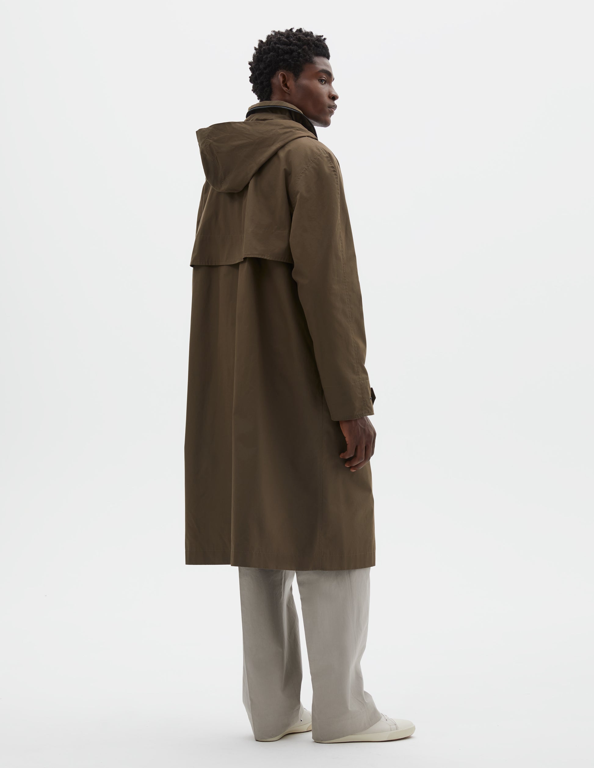 Full body image, back view of model wearing greenish brown ventile cotton poplin longline mac with deep storm vent and packable hood. Styled over beige trousers and off white canvas trainers.