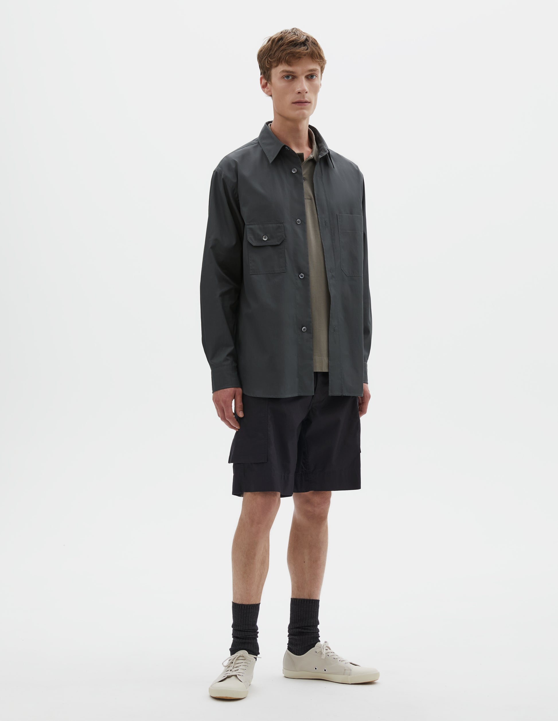 Full body image, front view of model wearing dark greenish blue long sleeve cotton shirt, with wide raised placket, asymmetrical patch pockets and mother of pearl buttons. Styled over green cotton polo shirt, with black cargo shorts, black socks and beige canvas trainers.