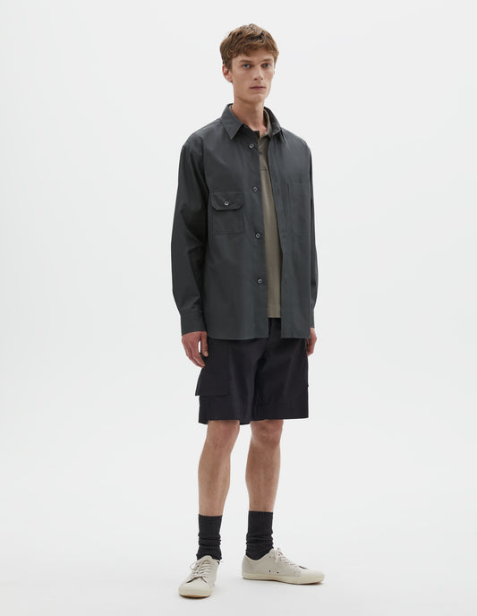 Full body image, front view of model wearing dark greenish blue long sleeve cotton shirt, with wide raised placket, asymmetrical patch pockets and mother of pearl buttons. Styled over green cotton polo shirt, with black cargo shorts, black socks and beige canvas trainers.