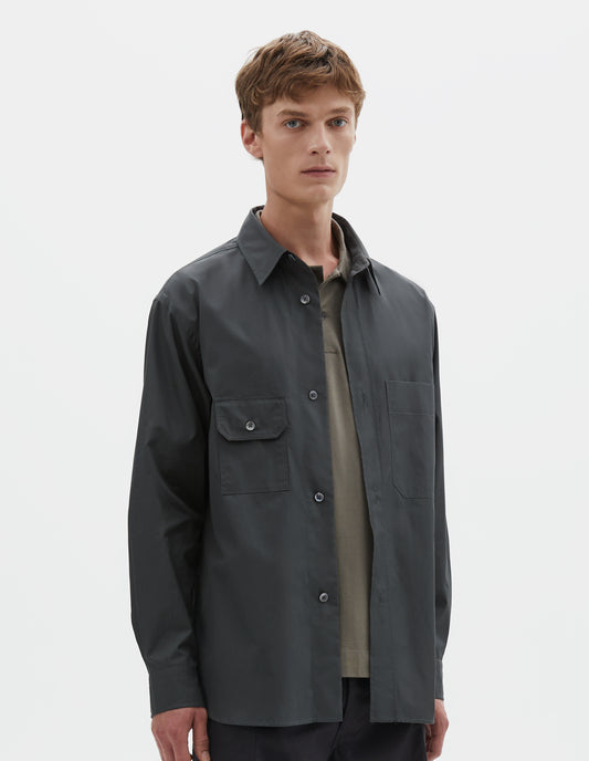 Cropped body image, front view of model wearing dark greenish blue long sleeve cotton shirt, with wide raised placket, asymmetrical patch pockets and mother of pearl buttons. Styled over green cotton polo shirt, with black cargo shorts.