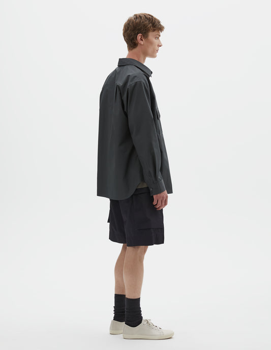 Full body image, back view of model wearing dark greenish blue long sleeve cotton shirt, with mother of pearl buttons cuffs and pleats on back yoke. Styled with black cargo shorts, black socks and beige canvas trainers.