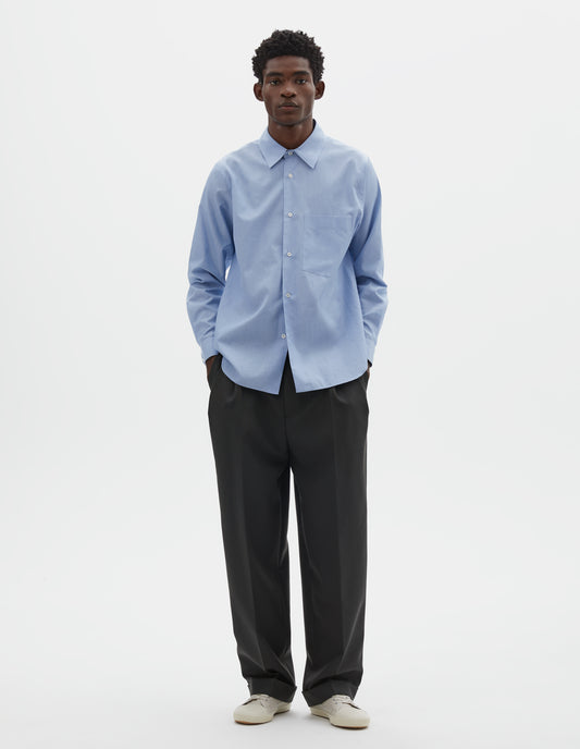 Full body image, front view of model wearing pale blue cotton shirt with chest pocket, curved hem and mother of pearl buttons. Styled with navy blue pleated trousers and beige trainers.