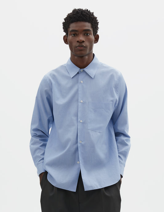 Cropped body image, front view of model wearing pale blue cotton shirt with chest pocket, curved hem and mother of pearl buttons. Styled with navy blue pleated trousers.