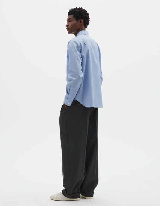 Full body image, back view of model wearing pale blue cotton shirt, with curved hem, pleats on back yoke and mother of pearl button cuffs. Styled with navy blue pleated trousers and beige trainers.