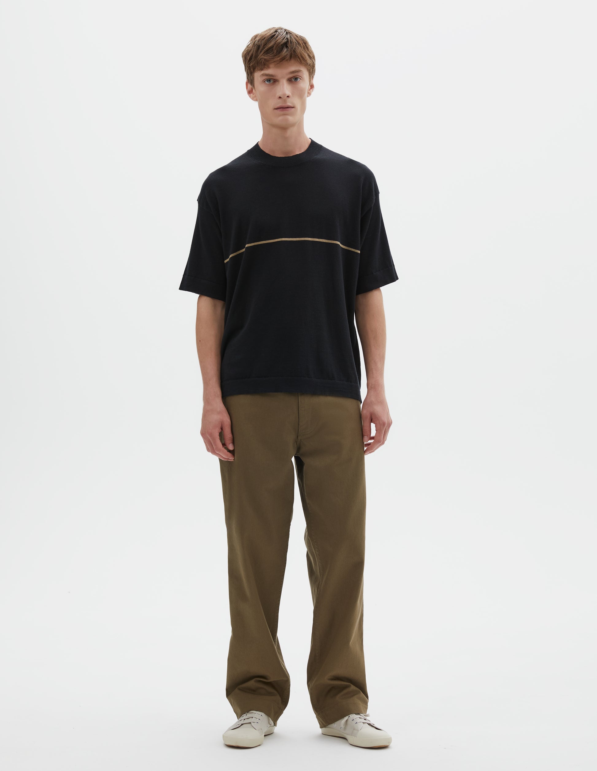Full body image, front view of model wearing regular, generous fit, black knitted t-shirt with deep rib crew neck, set in sleeves and single dark beige stripe across body. Styled with khaki green trousers and beige canvas trainers.