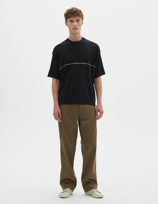 Full body image, front view of model wearing regular, generous fit, black knitted t-shirt with deep rib crew neck, set in sleeves and single dark beige stripe across body. Styled with khaki green trousers and beige canvas trainers.