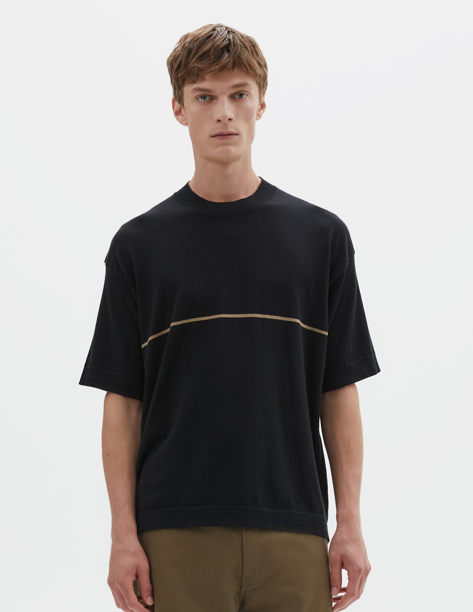 Cropped body image, front view of model wearing regular, generous fit, black knitted t-shirt with deep rib crew neck, set in sleeves and single dark beige stripe across body. Styled with khaki green trousers.