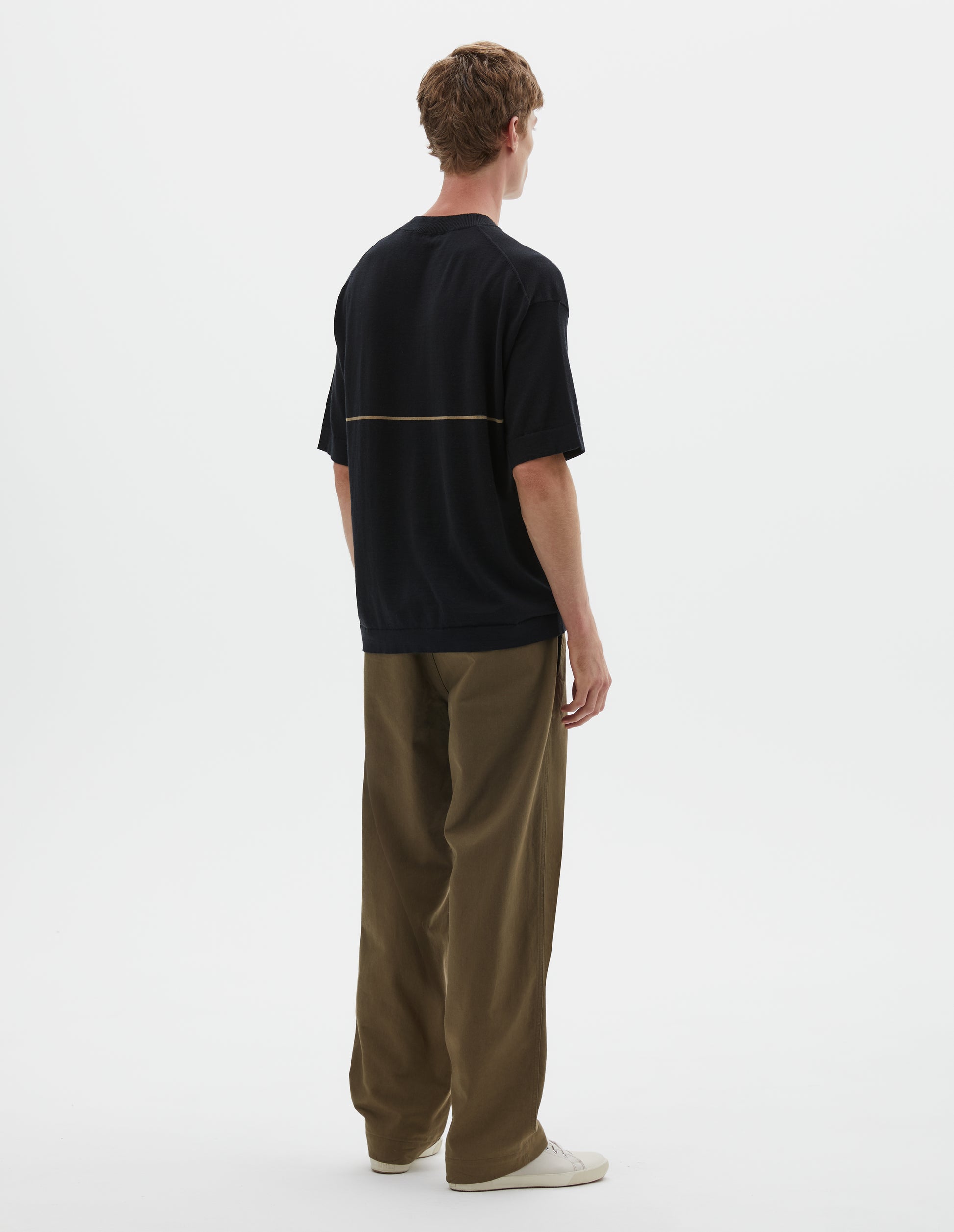 Full body image, back view of model wearing regular, generous fit, black knitted t-shirt with deep rib crew neck, set in sleeves and single dark beige stripe across body. Styled with khaki green trousers and beige canvas trainers.