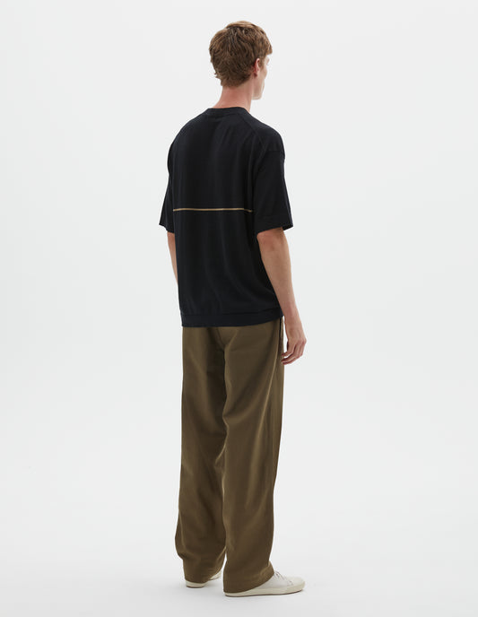 Full body image, back view of model wearing regular, generous fit, black knitted t-shirt with deep rib crew neck, set in sleeves and single dark beige stripe across body. Styled with khaki green trousers and beige canvas trainers.