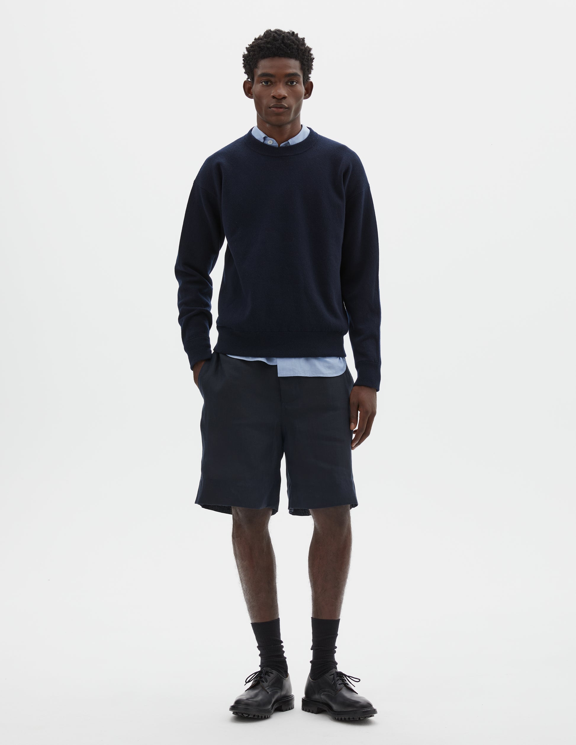 Full body image, front view of model wearing relaxed  dark blue knitted sweatshirt with rib collar, cuffs and hem. Styled over light blue shirt, with dark blue shorts and black socks with black leather shoes.