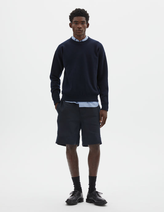Full body image, front view of model wearing relaxed  dark blue knitted sweatshirt with rib collar, cuffs and hem. Styled over light blue shirt, with dark blue shorts and black socks with black leather shoes.