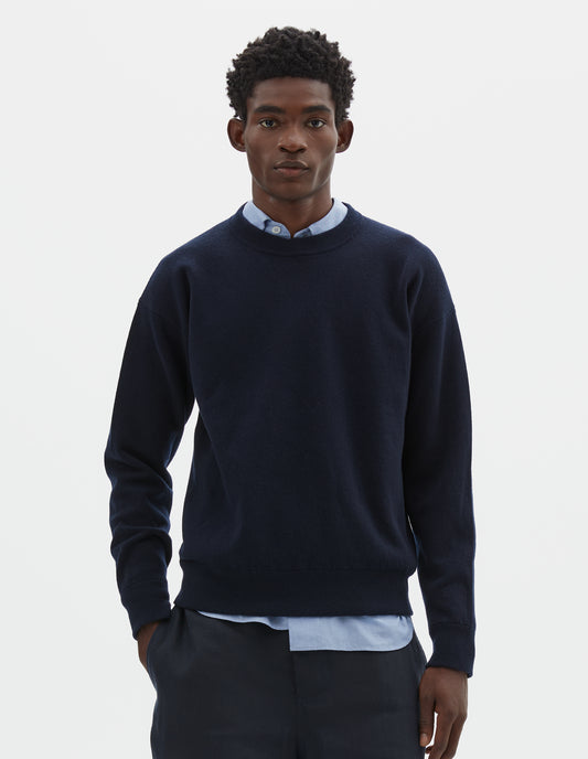 Cropped body image, front view of model wearing relaxed dark blue knitted sweatshirt with rib collar, cuffs and hem. Styled over light blue shirt and with dark blue shorts.