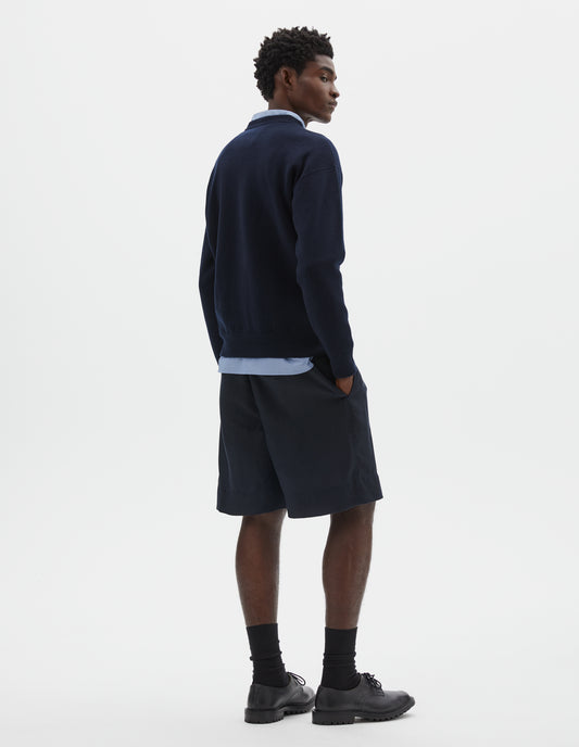 Full body image, back view of model wearing relaxed  dark blue knitted sweatshirt with rib collar, cuffs and hem. Styled over light blue shirt, with dark blue shorts and black socks with black leather shoes.