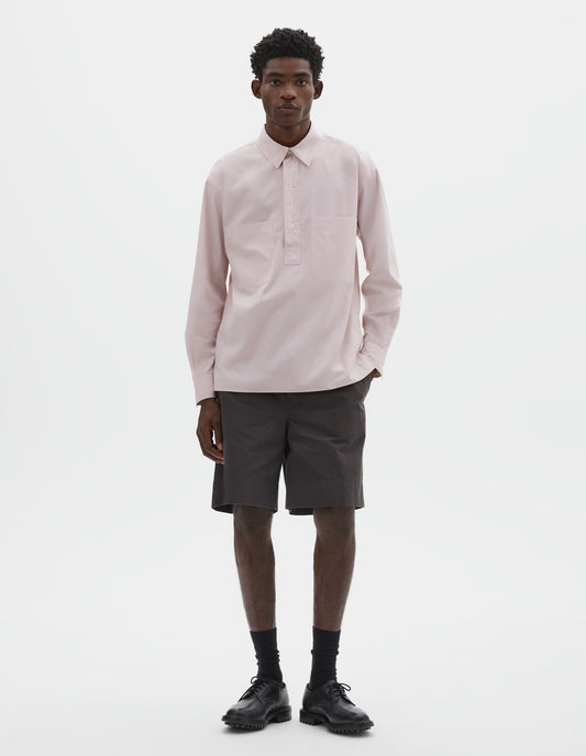 Full body image, front view of model wearing pink cotton long sleeve shirt with stitch through raised placket, front patch pockets and deep side split vents. Styled with brown shorts, black socks and black leather brogues.