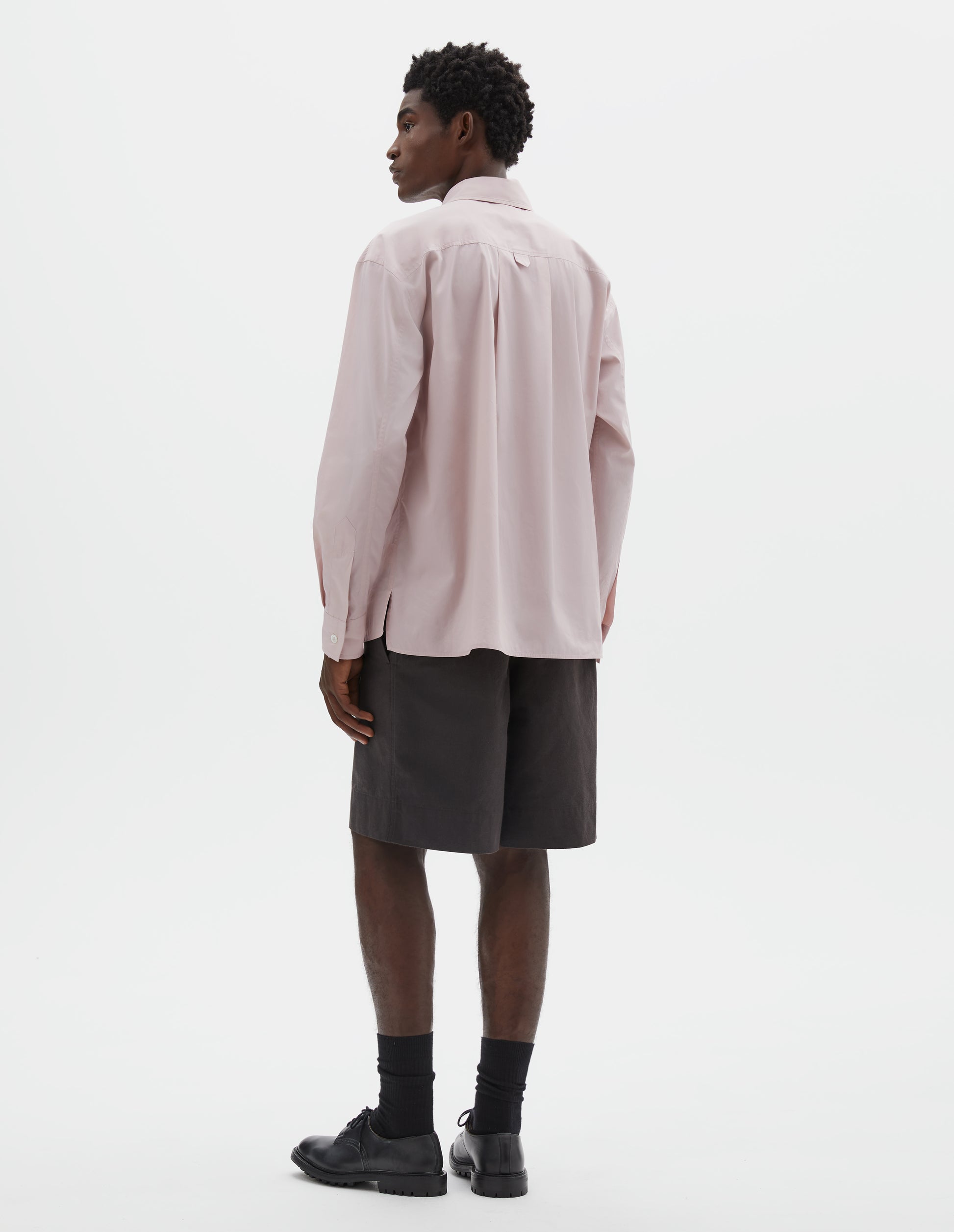 Full body image, back view of model wearing pink cotton long sleeve shirt with deep side split vents, elongated back and box pleat on back yoke. Styled with brown shorts, black socks and black leather brogues.