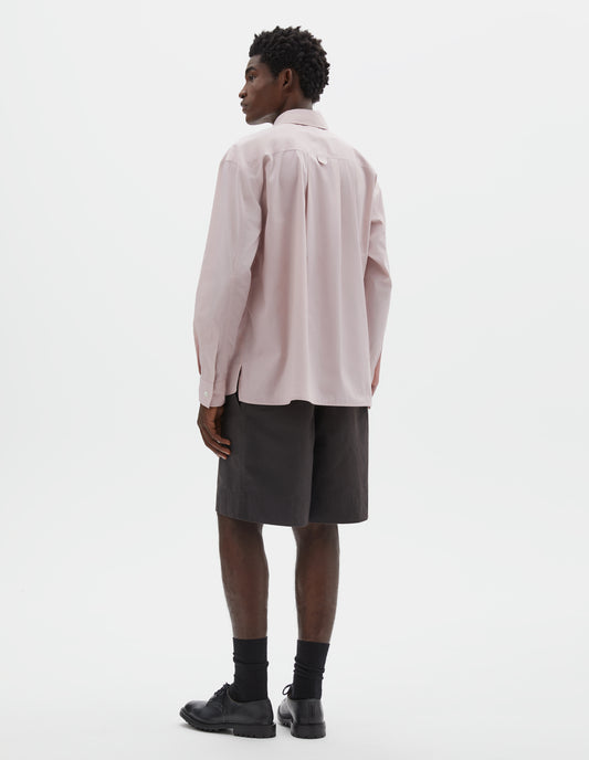 Full body image, back view of model wearing pink cotton long sleeve shirt with deep side split vents, elongated back and box pleat on back yoke. Styled with brown shorts, black socks and black leather brogues.
