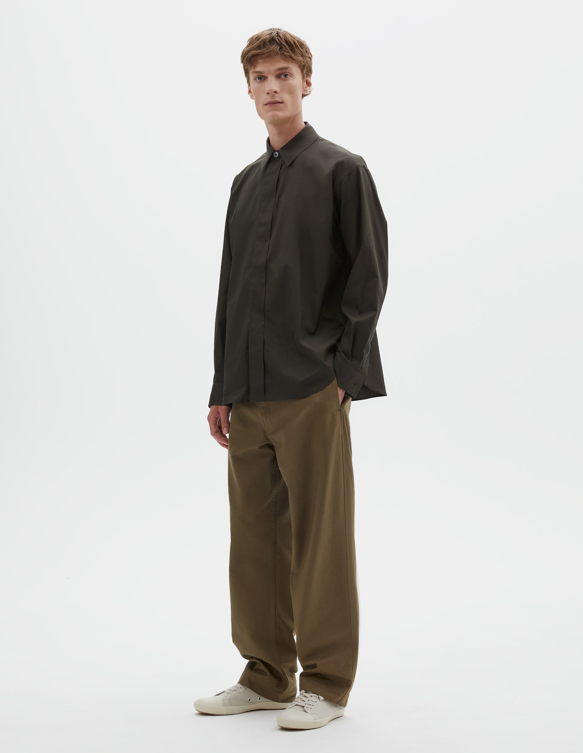 Full body image, front view of model wearing dark green cotton poplin long sleeve shirt with concealed button placket and curved hem. Styled with light green trousers and beige canvas trainers.