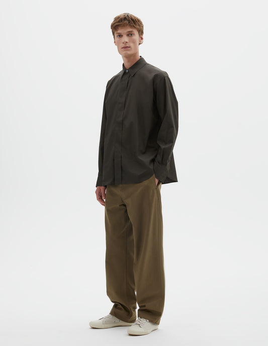 Full body image, front view of model wearing dark green cotton poplin long sleeve shirt with concealed button placket and curved hem. Styled with light green trousers and beige canvas trainers.