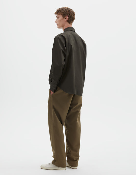 Full body image, back view of model wearing dark green cotton poplin long sleeve shirt, with horn button closure on cuffs and curved hem. Styled with light green trousers and beige canvas trainers.