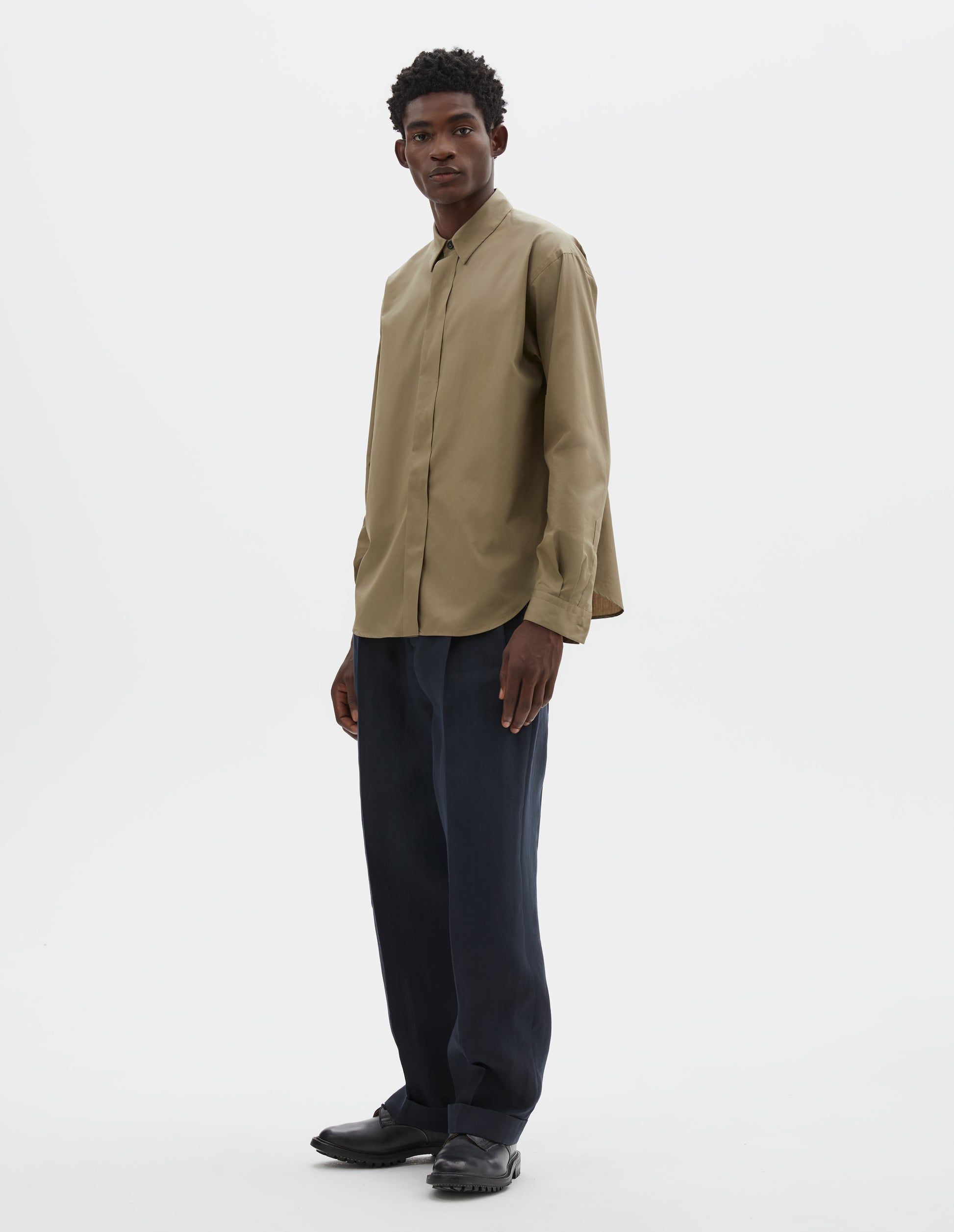 Full body image, front view of model wearing beige cotton poplin long sleeve shirt with concealed button placket and curved hem. Styled with dark navy trousers and black leather brogues.