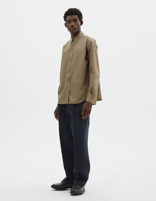 Full body image, front view of model wearing beige cotton poplin long sleeve shirt with concealed button placket and curved hem. Styled with dark navy trousers and black leather brogues.