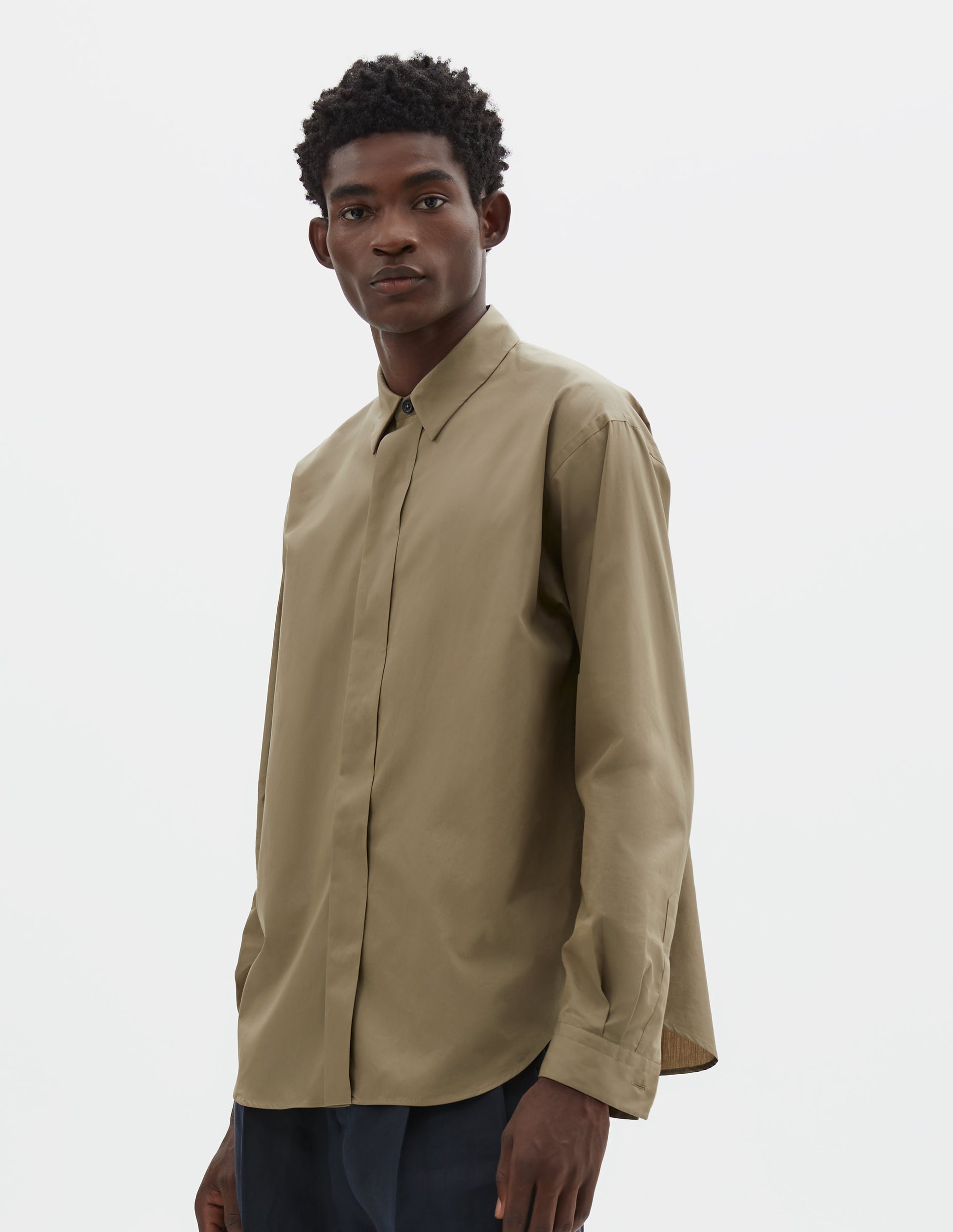 Cropped body image, front view of model wearing beige cotton poplin long sleeve shirt with concealed button placket and curved hem. Styled with dark navy trousers.