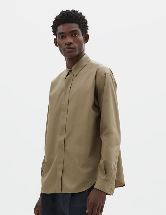 Cropped body image, front view of model wearing beige cotton poplin long sleeve shirt with concealed button placket and curved hem. Styled with dark navy trousers.
