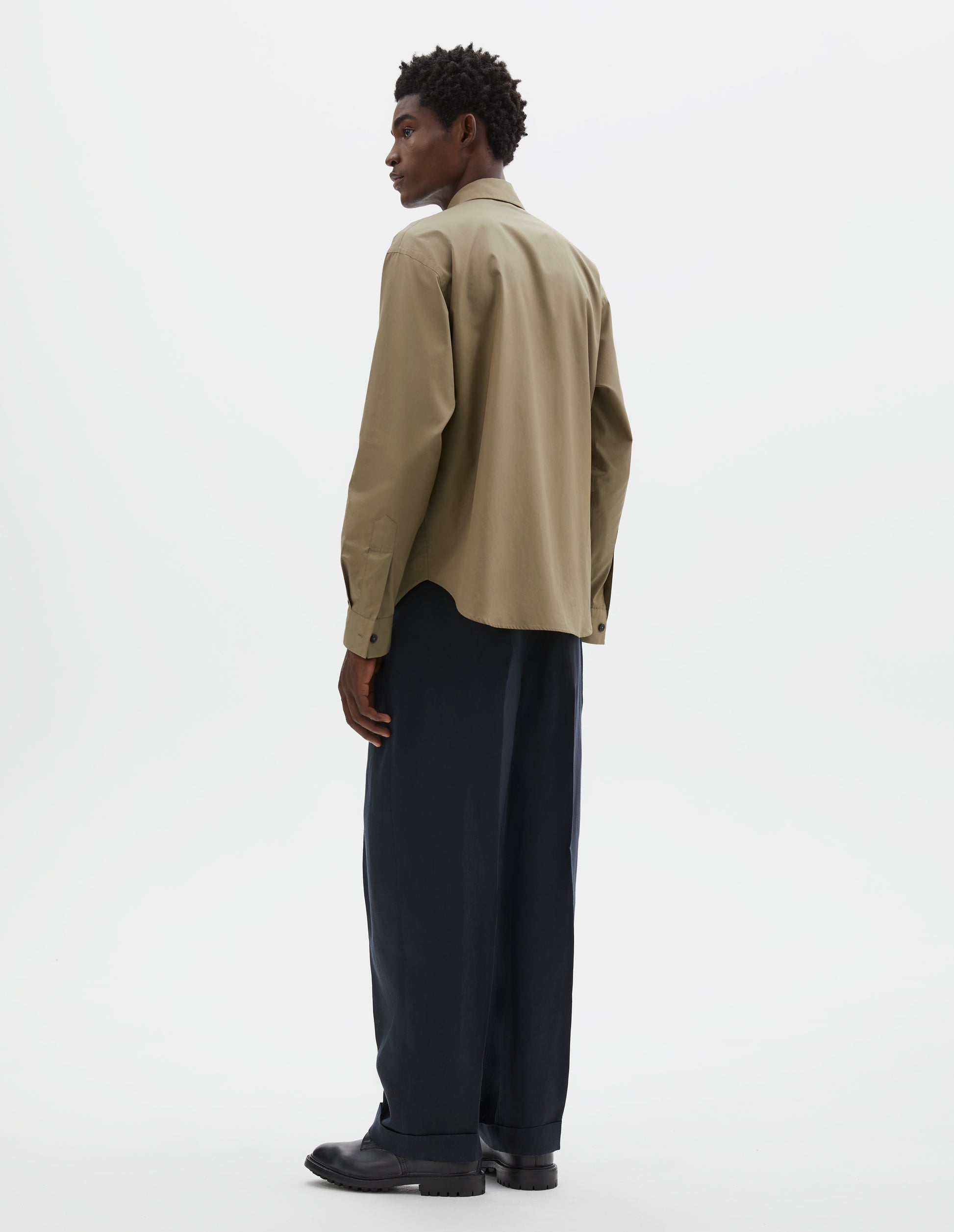 Full body image, back view of model wearing beige cotton poplin long sleeve shirt, with horn button closure on cuffs and curved hem. Styled with dark navy trousers and black leather brogues.