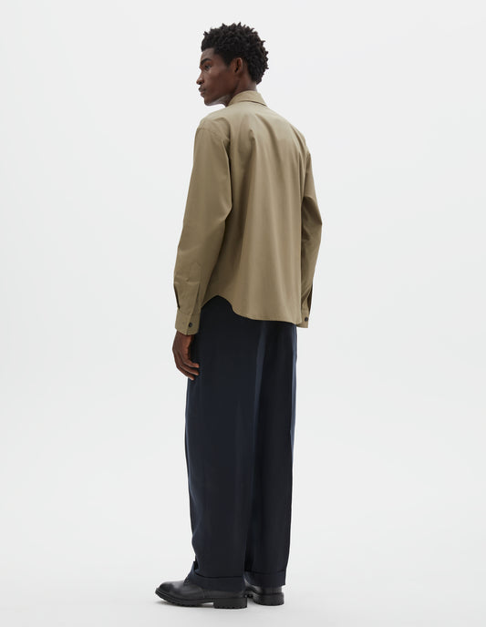 Full body image, back view of model wearing beige cotton poplin long sleeve shirt, with horn button closure on cuffs and curved hem. Styled with dark navy trousers and black leather brogues.