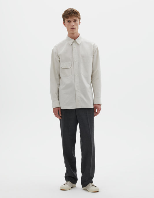 Full body image, front view of model wearing white long sleeve cotton shirt with patch pocket and button-through flap pocket on chest and raised placket with mother of pearl buttons. Styled with dark grey trousers and white canvas trainers.