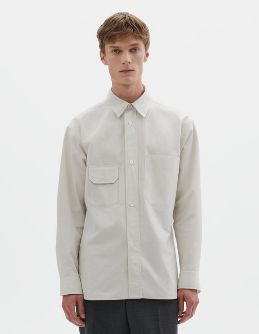 Cropped image, front view of model wearing white long sleeve cotton shirt with patch pocket and button-through flap pocket on chest and raised placket with mother of pearl buttons. Styled with dark grey trousers. 
