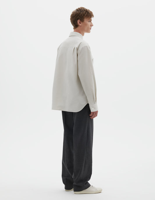 Full body image, back view of model wearing white long sleeve cotton shirt with mother of pearl buttons on cuffs and pleats on back yoke. Styled with dark grey trousers and white canvas trainers.