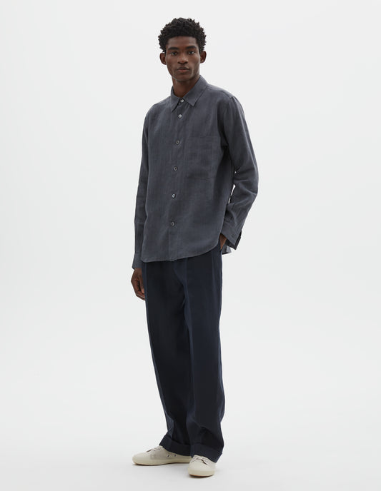 Full body image, front view of model wearing dark grey cotton long sleeve shirt, with mother of pearl buttons, curved hem and chest patch pocket. Styled with dark navy blue trousers and off white canvas trainers.