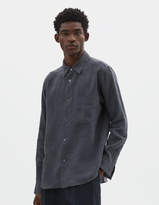Cropped body image, front view of model wearing dark grey cotton long sleeve shirt, with mother of pearl buttons, curved hem and chest patch pocket. Styled with dark navy blue trousers.